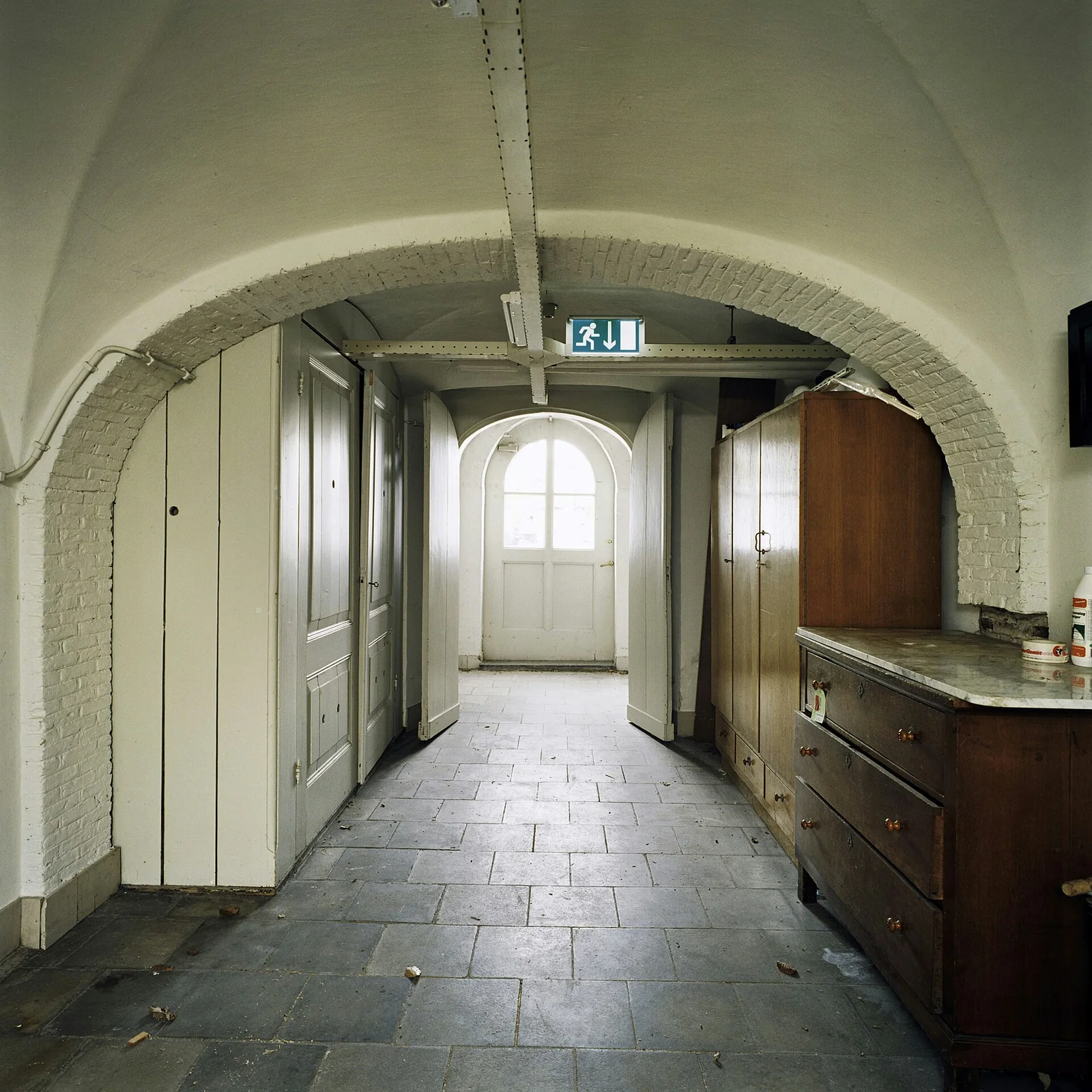 Een wit gepleisterde souterrainhal met gewelf, stenen vloer en daglicht.